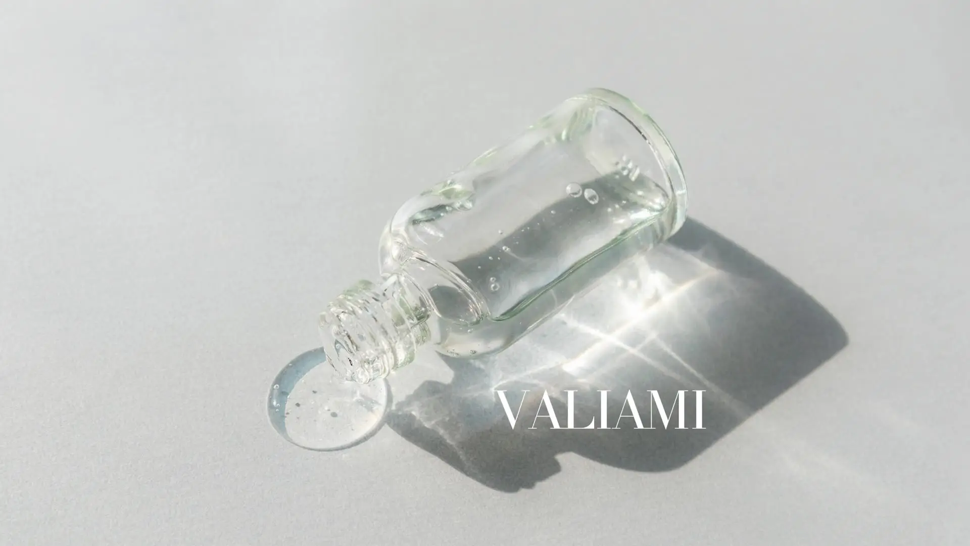 Flacon de soin VALIAMI en verre transparent, goutte de sérum et reflets de lumière sur fond gris clair